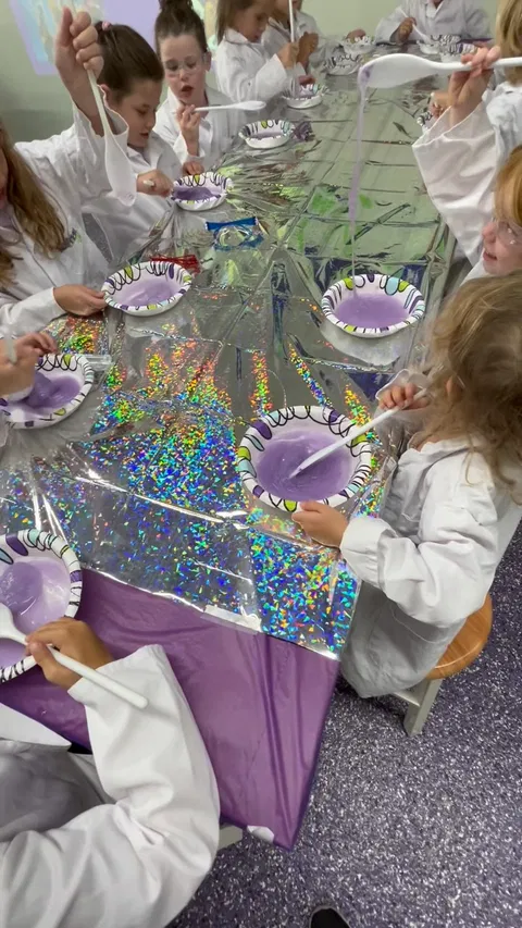 Children in lab coats making purple slime at a decorated table